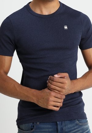Mand iført en tætsiddende mørkeblå t-shirt med korte ærmer og en lille hvid logo på øverste venstre bryst, kombineret med blå jeans.