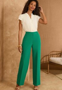 Pantaloni verdi a gamba larga con una vestibilità sartoriale, abbinati a una blusa crema con pieghe e un colletto sottile. Accento con un semplice bracciale d'oro.