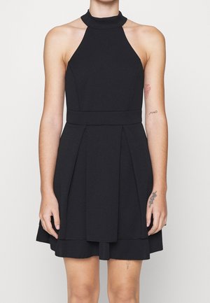 Femme portant une robe noire sans manches à col halter avec une jupe plissée, montrant des tatouages sur les deux bras et jambes, debout devant un fond uni.