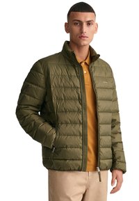 Chaqueta acolchada de color verde oliva con costuras horizontales, cuello alto y cremallera frontal. Llevada sobre una camisa polo de color mostaza. Diseño casual.