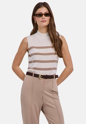 Woman wearing sleeveless beige and white striped top, high-waist beige pants with dark belt, and small brown sunglasses, standing with hands behind back.