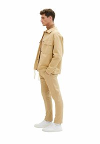 Beige jacka med fickor, i kombination med matchande beige byxor och vita sneakers. Mjuk textur och avslappnad passform, minimalistisk design.