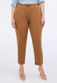 Pantaloni chinos in cotone marrone con vestibilità rilassata, orli arrotolati, tasche frontali e chiusura a bottone, abbinati a sandali con tacchi a cerchio trasparenti.