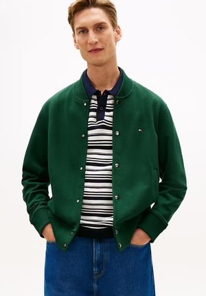 Giovane uomo che indossa una giacca verde con bottoni sopra una polo a righe bianche e nere con jeans blu, in piedi con le mani in tasca.