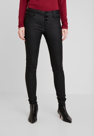 Femme portant un pantalon noir ajusté avec détail de boutons, des bottines noires pointues et un haut bordeaux à manches longues, debout sur un sol gris.