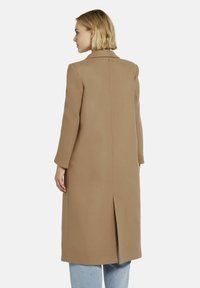 Langer, beiger Mantel mit Reverskragen, gerader Silhouette und Rückenbelüftung. Aus einem weichen Material gefertigt, mit minimalen Designdetails.