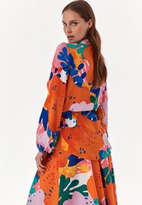 Färgglad blus med överdrivna ärmar och peplummidja, med abstrakta mönster i orange, rosa, blått och grönt på en ljus bakgrund.