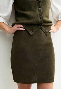 Falda de punto acanalado en verde oliva con forma ajustada, que presenta una cintura con botones y una textura sutil. Combinada con una blusa blanca.