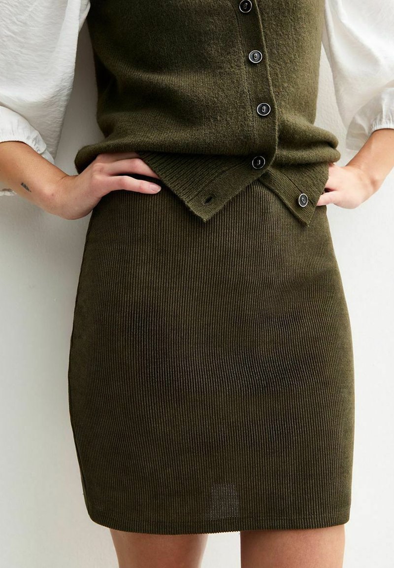 Falda de punto acanalado en verde oliva con forma ajustada, que presenta una cintura con botones y una textura sutil. Combinada con una blusa blanca.