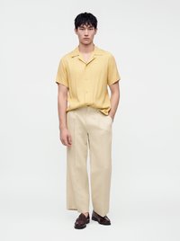 Jeune homme portant une chemise jaune à manches courtes, un pantalon beige à jambes larges et des mocassins marron foncé, debout avec une main dans la poche sur fond blanc.