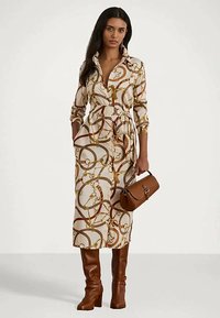 Mujer vistiendo un vestido midi con cinturón y estampado de cadenas y cuerdas, botas marrones hasta la rodilla y sosteniendo un bolso de cuero marrón.