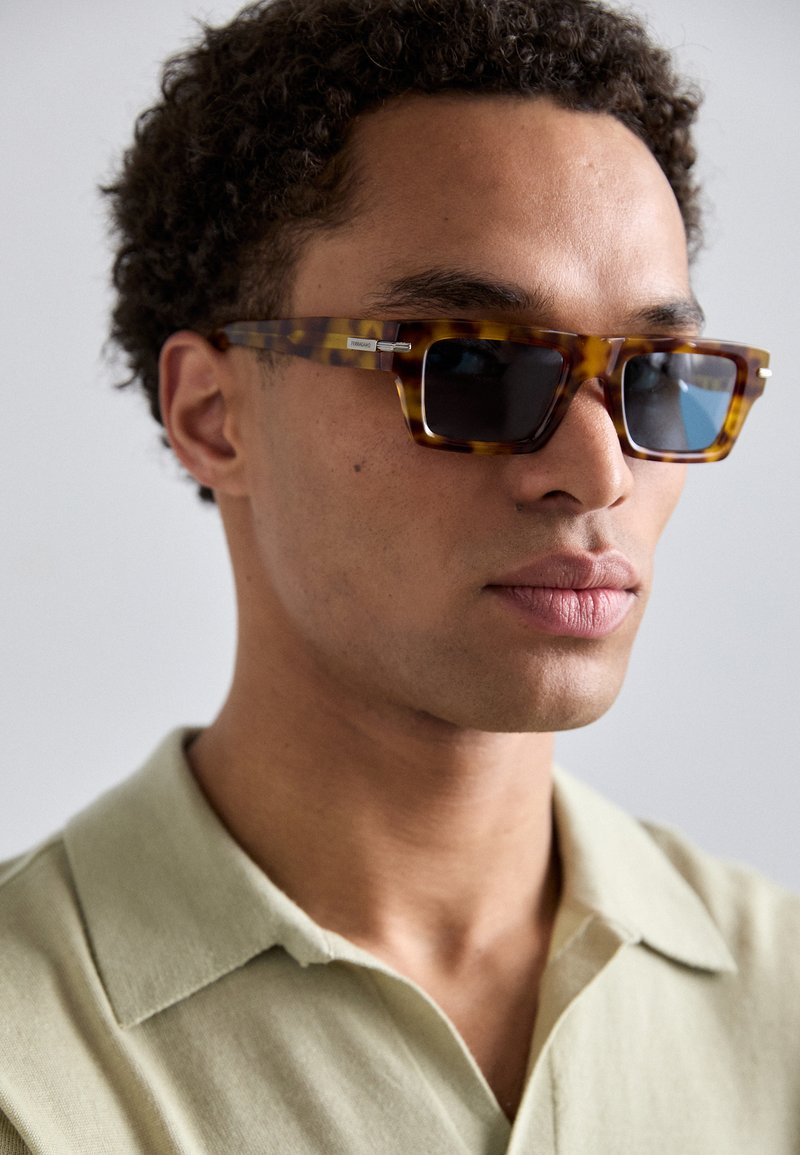 Óculos de sol quadrados em padrão tartaruga com lentes escuras, com detalhes em prata nas dobradiças, usados com uma camisa de colarinho verde claro. Superfície texturizada visível.