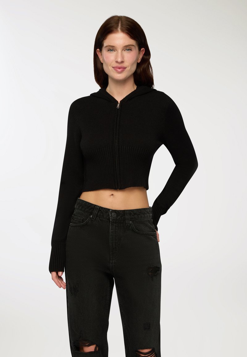 Crop top nero con zip e texture a coste, abbinato a jeans neri strappati, con vita alta e grandi strappi.