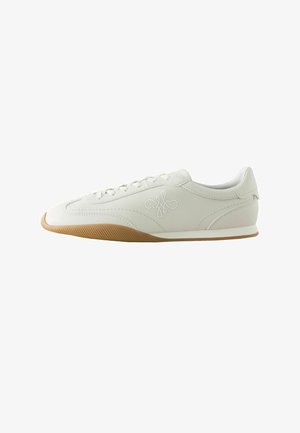 Hvid lav sneaker med brun gummisål, syningsdetaljer, snørelukning foran og broderet logo på siden.