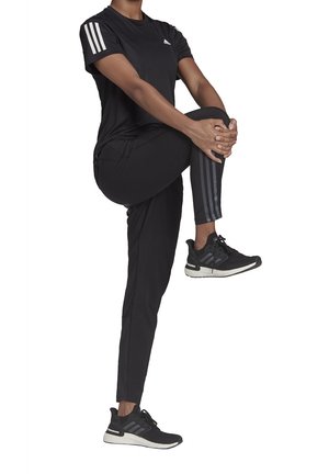 Černý sportovní outfit zahrnující krátký rukáv s třemi bílými pruhy a kalhoty s úzkým střihem. Nošeno s černými teniskami, odhalujícími texturované detaily.