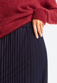 Burgundy knit sweater overlaps with a navy pleated skirt. Skirt features vertical pleats and a smooth waistband, with a hand resting on it.