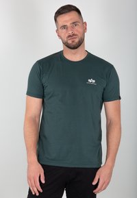 Dunkelgrünes Baumwoll-T-Shirt mit kurzen Ärmeln, Rundhalsausschnitt und einem kleinen weißen Logo auf der linken Brust. Weicher Stoff, lässige Passform.