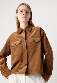 Femme aux longs cheveux portant des boucles d'oreilles créoles en argent et des lunettes de style aviateur transparentes, une veste en velours côtelé marron à boutons, et un pantalon blanc.