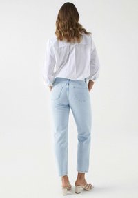 Jeans de mezclilla azul claro con corte recto, que cuentan con dos bolsillos traseros y una cintura alta, combinados con una camisa blanca de botón y sandalias.