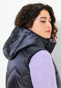 Marinblå puffväst med huva, med ett quiltat mönster. Tillverkad av glänsande, lätt material, lagerd över en lavendelfärgad ribbad tröja.