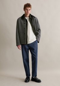 Mannen geruite jas in zwart en grijs met een donkergroene kraag, gedragen over een wit shirt en blauwe jeans, gecombineerd met zwarte schoenen.