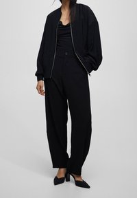 Zwarte bomberjack met ritssluiting, gedragen over een zwarte coltrui. Zwarte hoge taille, wijde broek en puntige hakken.