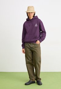 Sweatshirt violet avec poche avant et logo ; pantalon olive vert, jambes larges ; chaussures noires à enfiler ; chapeau seau beige ; fond plat.