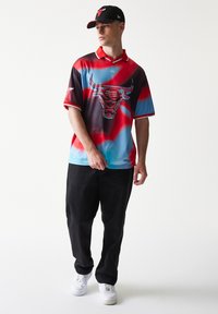 Πολύχρωμο πόλο με στάμπα βούβαλου, κυρίως κόκκινο, μπλε και μαύρο. Φορεμένο με μαύρο παντελόνι και καπέλο που ταιριάζει. Άσπρα αθλητικά παπούτσια.