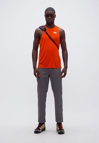 Άνδρας με πορτοκαλί αμάνικο πουκάμισο, γκρι παντελόνι, παπούτσια πεζοπορίας, γυαλιά ηλίου και μαύρη τσάντα χιαστί σε λευκό φόντο.