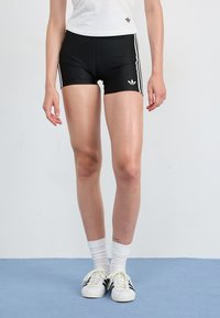 Calças de desporto pretas com listras brancas laterais, apresentando um pequeno logótipo. Usadas com uma blusa branca e ténis clássicos preto e branco.