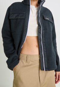 Kurtka polarowa w ciemnoszarym kolorze z czarnymi akcentami, z wysokim kołnierzem, dwoma kieszeniami na piersi i zamkiem błyskawicznym z przodu. Noszona na krótki top.