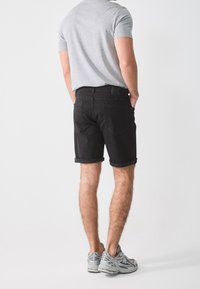 Pantalones cortos de mezclilla negra con dobladillos enrollados, presentados con bolsillos traseros y una camiseta ajustada gris. Zapatillas deportivas informales completan el look.