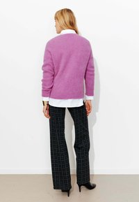 Femme de dos portant un pull lavande sur une chemise blanche, un pantalon large à carreaux noirs, des talons noirs et un bracelet manchette en or.