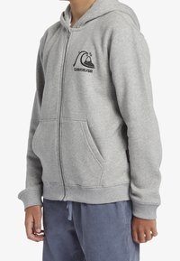 Persona che indossa una felpa grigia chiaro con zip, con un logo di onda e montagna nera sul petto, abbinata a pantaloni blu con cordino.