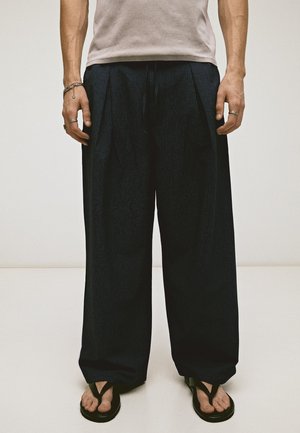 Personne portant un pantalon noir ample, un haut beige clair, des sandales noires et plusieurs bagues et bracelets en argent, debout devant un fond uni.