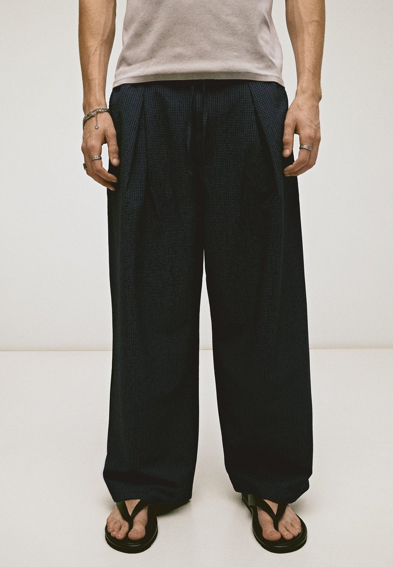 Personne portant un pantalon noir ample, un haut beige clair, des sandales noires et plusieurs bagues et bracelets en argent, debout devant un fond uni.