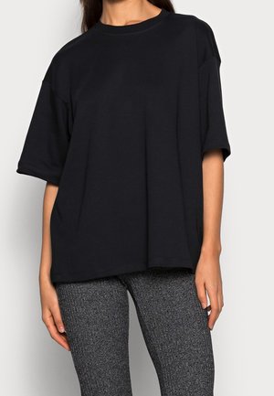 Vrouw die een losse zwarte T-shirt met korte mouwen en nauwsluitende grijze, getextureerde broek draagt, tegen een effen achtergrond.