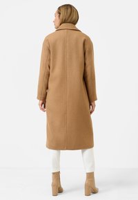 Lange, camelkleurige wollen jas met een rechte snit, brede kraag en zijzakken. Model draagt witte broek en beige hakken.