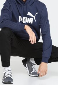 Felpa navy con logo "PUMA" bianco, abbinata a pantaloni della tuta neri e scarpe da ginnastica in rete grigia, con lacci e dettagli bianchi.