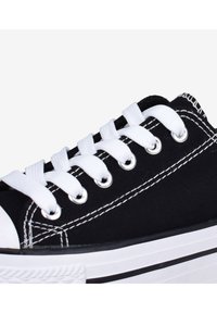 Sneaker nera in tela con lacci bianchi, occhielli in metallo e punta in gomma bianca. Cuciture bianche notevoli lungo i lati.