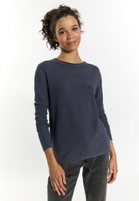 Top din tricotaje cu model waffles de culoare albastru marin, cu maneci lungi, decolteu rotund și mansete rulate, asociat cu blugi din denim gri închis. Textură moale și croi lejer.