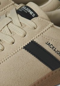 Sneaker en suède beige clair avec une bande de caoutchouc noir. Lacets tissés plats, détails cousus et semelle en caoutchouc texturé. Étiquette de marque visible.