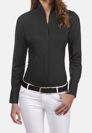 Vrouw die een nauwsluitend zwart shirt met lange mouwen en een opstaande kraag draagt, witte broek en een zwarte riem met een zilverkleurige gesp.