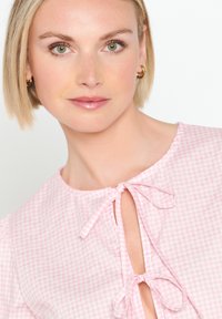 Blusa in gingham rosa con scollatura rotonda, chiusure a fiocco davanti e maniche a sbuffo. Tessuto in cotone morbido con un motivo a scacchi.