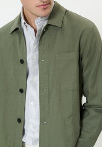 Muž nosiaci zelenú zapínaciu bundu cez pruhovanú bielo-sivú košeľu, zobrazený od horného trupu a časť tváre otočená do strany.