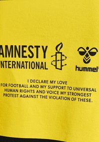 Gul bomullsskjorta med svart text och logotyper för Amnesty International och hummel, som uttrycker stöd för mänskliga rättigheter och fotboll.