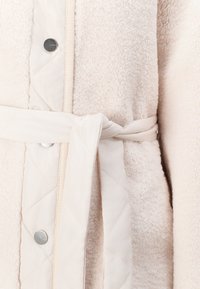 Beige fleece robe with quilted light beige accents, featuring a tie belt and silver snap buttons for closure. Soft textured fabric.