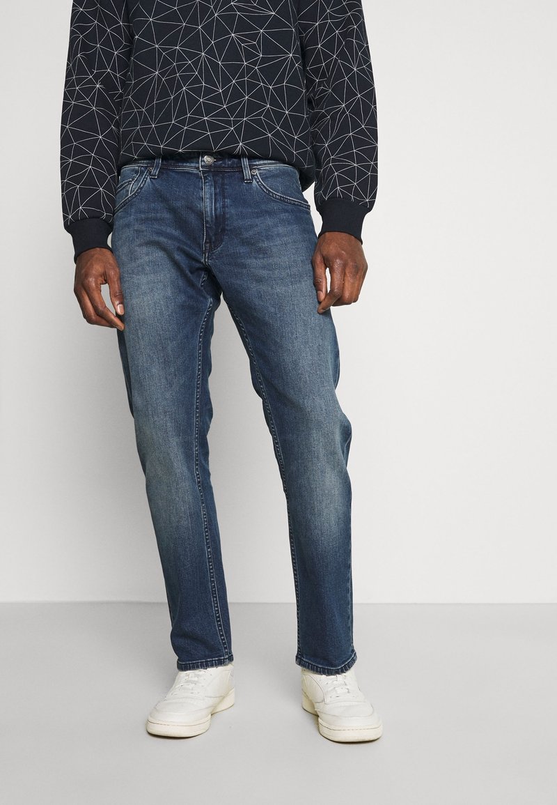 Dark blue denim jeans with a straight-leg cut, featuring faded sections and five-pocket design. Worn with a patterned navy sweatshirt and white sneakers.