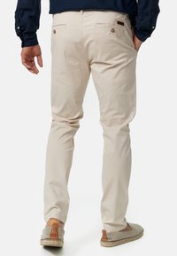 Pantalon en coton beige clair avec poches arrière, détails à boutons et coupe ajustée. Porté avec des espadrilles grises.