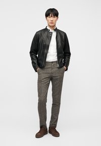 Mladý muž stojaci s rukami v kapse, oblečený v čiernej koženej bunde, bielej košeli, sivých nohaviciach a hnedých teniskách na jednoduchom pozadí.